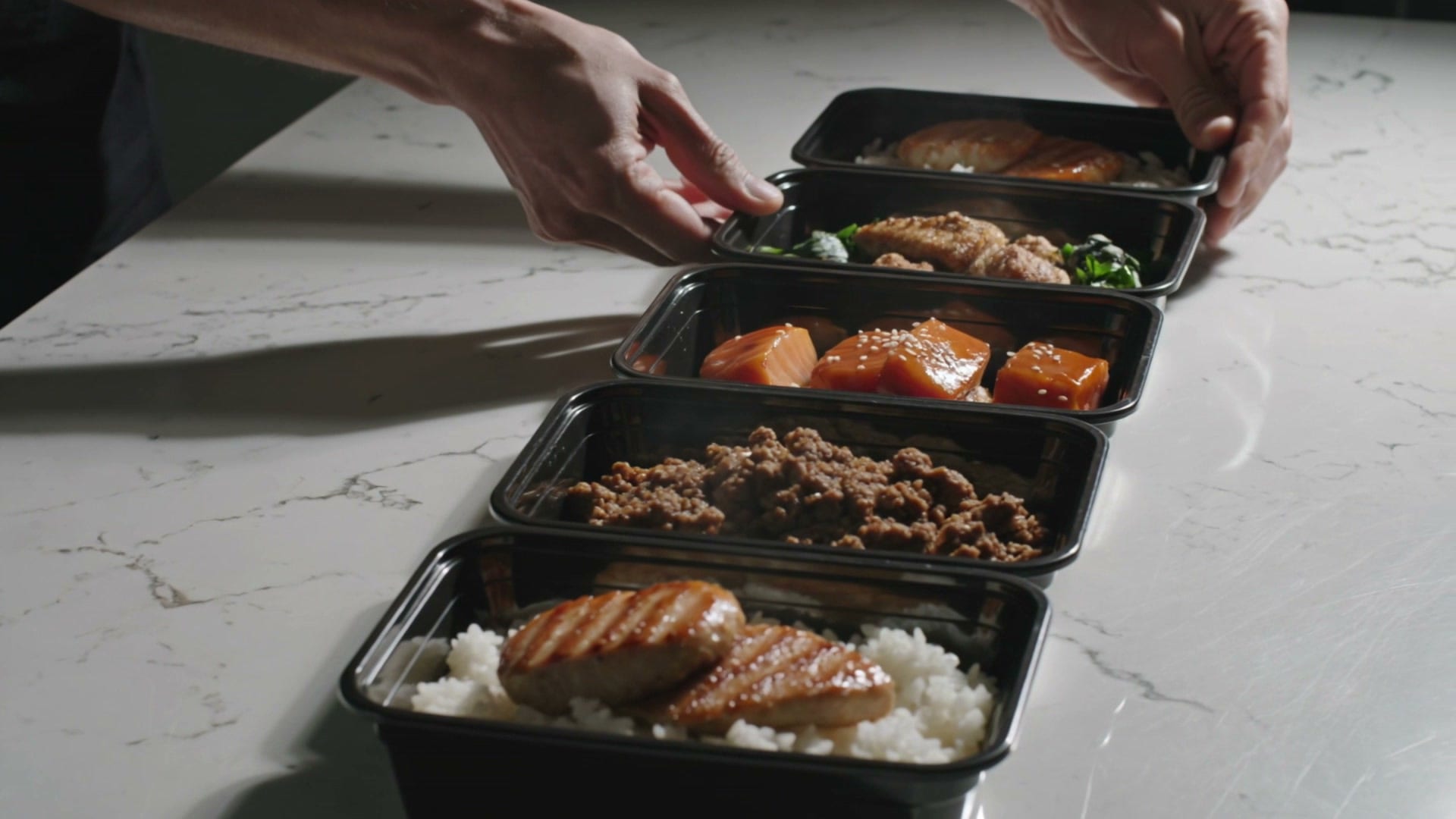 Five meal-prep containers of chicken, salmon and ground beef over rice, lined up on a marble counter